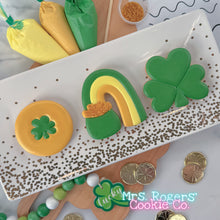 Load image into Gallery viewer, An overhead view of three decorated cookies on a white rectangular platter with gold polka dots. From left to right: a round gold coin cookie with a green shamrock in the center, the rainbow-into-pot-of-gold cookie, and a large green three-leaf shamrock cookie. Icing bags in yellow and green, gold sprinkles, and plastic gold coins are scattered around the platter on a white marble surface.