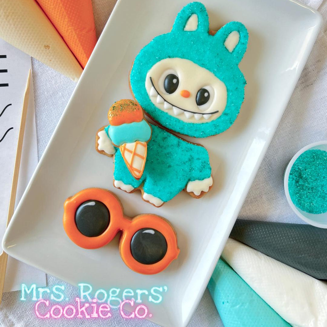 Cookie decorated like a character in a blue costume with orange glasses on a white plate, with 'Mrs. Rogers' Cookie Co.