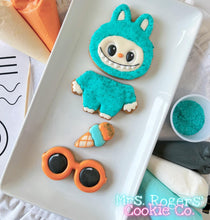 Load image into Gallery viewer, Cookie shaped like a character in a blue outfit with sunglasses on a white plate, branded &#39;Mrs. Rogers&#39; Cookie Co&#39;.
