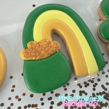 Load image into Gallery viewer, A close-up of a sugar cookie shaped like a rainbow ending in a pot of gold. The rainbow has three stripes in dark green, light yellow, and golden orange. The "pot" at the bottom is dark green, topped with a thick layer of orange icing and sparkling gold sugar crystals to represent overflowing gold coins.
