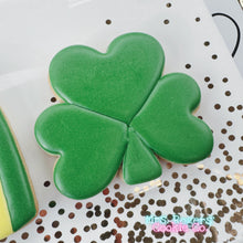Load image into Gallery viewer, A close-up of a large sugar cookie shaped like a three-leaf shamrock. The cookie is covered in smooth, vibrant green royal icing with subtle indentations defining the three heart-shaped leaves and the small stem at the bottom. It sits on a white plate decorated with metallic gold confetti.
